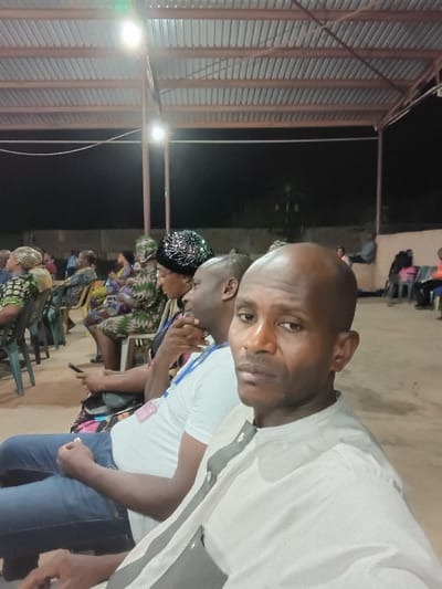 A bald man in a light-colored shirt is at the forefront of the image. Behind him, a person in a white t-shirt is visible. Further back, people are seated, some wearing patterned clothing and head coverings. The setting appears to be an outdoor event or gathering under a roofed structure with corrugated metal roofing. There are chairs visible throughout the frame, and the lighting suggests the photo was taken in the evening.