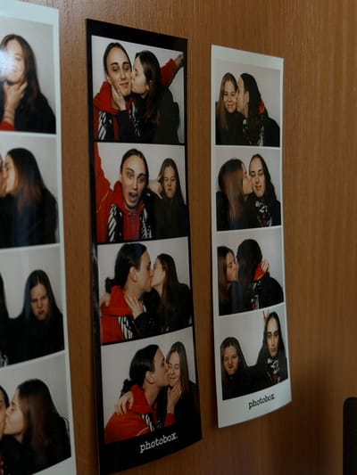 On a wooden surface, three photobooth strips are displayed. Two strips are fully visible, and a portion of the third strip is seen at the bottom of the frame. The strips are rectangular and feature multiple sequential images of two individuals, presumably a couple. The individuals are depicted in close proximity, engaged in affectionate poses such as kissing and embracing. The strips have a black border and include the word "photobox" printed on the side. The images are predominantly in black and white, though some strips show color variations due to lighting or film processing. The surface appears to be a wooden table, with a neutral tone and visible wood grain.