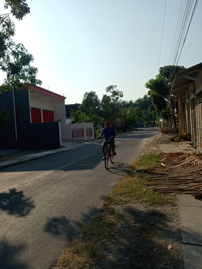 An overhead, eye-level shot captures a person riding a bicycle down a paved road in Tegalkodo, Indonesia. The sky is a clear, bright blue with the sun high in the sky, casting sharp shadows on the road and surrounding areas. To the left of the road, a modern house with dark grey and white walls and a red trim is visible. The house has two large red shutters. Adjacent to the house is a white fence with red bars. Across the street from the house, a sidewalk is lined with dry palm fronds. Beyond the houses, lush green trees and vegetation dot the landscape, with a small hill visible in the distance. The road is mostly empty, with the exception of the cyclist and a few distant motorcycles. The scene suggests a quiet, sunny day in a residential area.