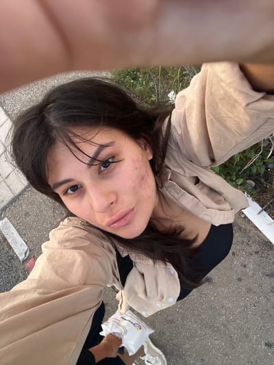 A young woman with dark hair and light skin is taking a selfie outdoors. She is wearing a beige open shirt over a black top. Her face is turned towards the camera, and she has some acne on her cheeks. She is holding a white object with colorful stripes in her right hand. The background shows a sidewalk with concrete slabs, asphalt, some green plants, and dry branches. The lighting suggests it is daytime. The presence of the sidewalk and the general urban environment, combined with the specific geographical reference to Kiryat Motzkin, Israel, indicates this scene is located in a public outdoor space within that city.