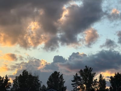 This media file shows a cloudy sky over a silhouette of trees and rooftops. The clouds are dramatic, with some areas illuminated by the sun in hues of orange and yellow, while others are dark gray. This suggests it is either sunrise or sunset. The light filtering through the clouds creates a dramatic effect, highlighting the edges of some clouds with a bright glow. The trees are dark and appear to be deciduous with some evergreen pines mixed in, their branches silhouetted against the vibrant sky. The rooftops are barely visible at the bottom, suggesting a rural or suburban setting. The overall mood is serene and awe-inspiring, capturing the beauty of a dynamic sky. The location context indicates this scene is likely in Грузино-3 Снт, Russia. There is no visible text in the image.