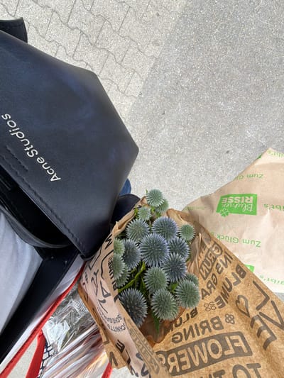 The image is a medium-angle, eye-level shot taken outdoors on a paved surface, likely a sidewalk or plaza. In the foreground, a bouquet of spiky, blue-green thistle-like flowers is wrapped in brown paper. The paper has a repeating pattern of text and graphics, including the brand name "BLUMEN RISSE" in green and "Zum Glück" in green. The text is partially obscured and some of it is upside down. To the left of the flowers is a black leather bag with "Acne Studios" embossed in silver on its surface. The bag appears to be a tote or shoulder bag. White and red fabric, along with a clear plastic-wrapped item, are visible beneath the bag. The background shows a light grey, textured pavement with visible seams between the paving stones. The overall impression is that of someone carrying shopping, including a floral purchase.
