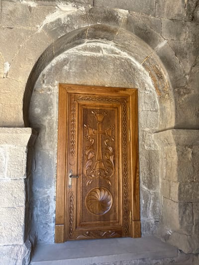 A rectangular, ornately carved wooden panel is set within a stone wall. The panel features a detailed depiction of a cross and a circular floral design, possibly a rose. The stone wall surrounding the panel has an arched top and is composed of rough-hewn blocks of grey stone, indicating an old structure. A metal latch can be seen on the panel.