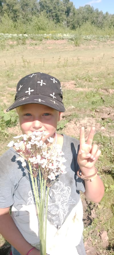 A young child wearing a black baseball cap with a white cross pattern and a gray t-shirt is holding a bouquet of small white flowers. The child is looking at the camera with a slight smile and holding up two fingers in a peace sign with their right hand. They are also wearing a braided bracelet on their right wrist and a pink necklace. The background appears to be an outdoor, rural setting with dry grass, dirt, and trees in the distance. The image was taken outdoors in Январцев ауылдық округі, Kazakhstan.