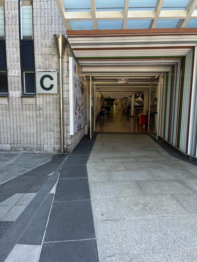 The image presents a striking architectural view, taken from a high vantage point looking down into a long, narrow covered walkway or atrium. The perspective creates a strong sense of depth, with the passage receding sharply into the distance.
On the left, the exterior of a building features large, gridded windows with light-colored frames, suggesting an adjacent outdoor space or another building section. Below the windows, a light-colored brick wall is visible, on which a white square sign displays a prominent black letter "C". Further down, several dark, rectangular window-like panels are integrated into the brickwork, and a metallic railing runs horizontally.
The central and most prominent feature is the elongated corridor. Its walls are distinctive, lined with vertical stripes in varying shades of white, brown, green, and grey. The floor of this passage in the foreground is adorned with a colorful, abstract pattern or artwork. As the corridor extends, it appears to transition into an indoor space, brightly illuminated by artificial lights, revealing what seem to be office areas or classrooms with furniture in the far distance. Horizontal metal railings parallel the length of this walkway.
To the right, a substantial wall composed of grey tiles forms the boundary of the viewing area. This wall is vertically divided into two sections: an upper part constructed from light grey, speckled granite-like tiles, and a lower section made of darker grey, solid-colored tiles.
The scene depicts a modern institutional or public building, aligning with a university or campus setting in Kelvin Grove, Australia. The mixture of natural light entering from the left and artificial lighting within the corridor suggests a thoughtfully designed covered or semi-indoor architectural space. No people are visible, indicating a quiet period. The most notable visible text is the "C" logo on the brick wall.