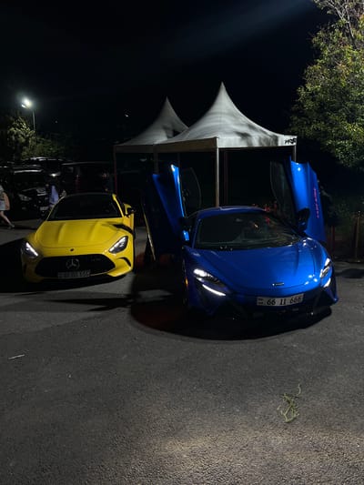 The image captures a scene at night in Jrvej, Armenia, featuring two prominent cars and a tent structure. A blue McLaren is positioned near the tent, with its doors open, revealing a partial view of the interior. Adjacent to the McLaren is a yellow Mercedes-AMG GT. Both vehicles have Armenian license plates, with the McLaren’s reading “66 II 999” and the Mercedes’ reading “66 BB 666”.

In the background, a white tent structure stands, partially obscured by tree foliage. A streetlight illuminates the scene. Several people are partially visible near a black vehicle, suggesting an event or gathering. The ground is paved.