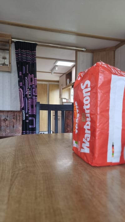 A low-angle, medium shot captures a loaf of Warburtons bread, prominently displayed in its red and white packaging, resting on a wooden table in what appears to be a mobile home or caravan. The bread bag is positioned towards the right side of the frame, with its text largely visible. In the background, a dark wooden chair is situated behind the table, and beyond that, a black and purple patterned curtain hangs. Further in the background, the interior of the mobile home is visible, including a skylight in a ceiling, kitchen cabinets, and what seems to be a window. The overall scene suggests a domestic setting, possibly a dining area within a caravan, located in Auchenblae, United Kingdom.