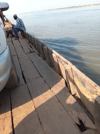 The image displays a wooden ferry traversing the Benue River in Ibi, Nigeria. In the foreground, the front left quarter of a silver vehicle is parked on the ferry's plank deck. An adult male, bald and wearing a blue and white checkered shirt with blue jeans, sits on the ferry's worn wooden railing, facing the open water. Partial view of another individual's legs in brown trousers is visible near the vehicle. The calm, wide river stretches into the distance under a clear sky, with a faint horizon line marking the opposite bank. Strong sunlight casts diagonal shadows across the wooden deck.