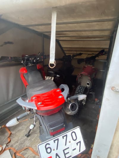 The image shows two motorcycles secured inside an enclosed trailer. One motorcycle is oriented towards the viewer and is partly red and gray. A white license plate is mounted on the rear, partially obscured, and contains digits "6.7." The license plate also contains "KE-7." A white strap with a D-ring is visible, secured to the front portion of the motorcycle. Another strap is visible securing another motorcycle. The trailer interior is constructed of plywood panels, and metal support beams are also visible. The location is Азёрскі сельскі Савет, Belarus.