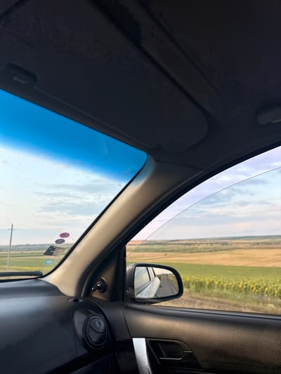 The image is an interior view from a car traveling through Carabetovca, Moldova. The perspective looks towards the windshield and out the passenger side window. In the foreground, the dark textured dashboard and door panel of the car are visible, along with a side mirror reflecting the road behind. Outside the car, a landscape of agricultural fields, including a section of sunflowers, stretches towards the horizon under a cloudy sky. The sky has a blue tint, likely from the windshield. A telephone pole is visible in the distance to the left. Several stickers are affixed to the windshield. There are no people visible in the image.