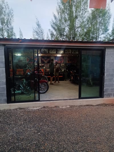 The image shows the exterior of a building in Surat Thani City Municipality, Thailand. The building is constructed of cinder blocks, with a wide opening containing sliding glass doors. Inside the building, several motorcycles are visible, along with chairs, framed pictures, and flags displayed on the back wall. Two people are standing inside the building. The ground outside the building is covered in small gravel. Trees are visible in the background. The sky is overcast.