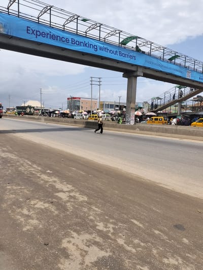 The image shows a wide, multi-lane highway in what appears to be a bustling urban environment. A person is seen crossing the road, seemingly against traffic. Above the road, a pedestrian bridge is adorned with a large advertisement for "Parallex Bank", and people are seen walking across the bridge. On the opposite side of the road, there's an active roadside market with vendors under umbrellas, and various vehicles including yellow taxis, a truck, and a few vans. The road surface in the foreground is worn and has patches of discoloration. Electrical poles and buildings are in the background, giving context to the scene. This scene is in Ibafo, Nigeria.