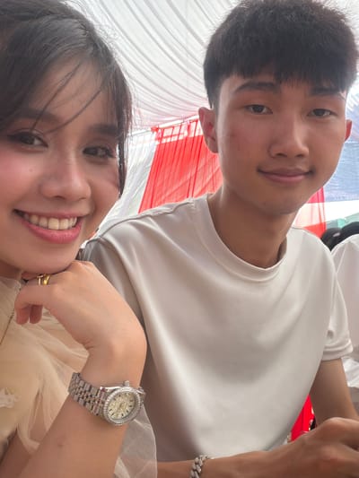 A close-up, high-angle photograph captures two young adults, a male and a female, in what appears to be a festive outdoor or semi-outdoor setting in Tay Son Commune, Vietnam. The image is a selfie, taken from below, looking upwards.
In the foreground, a female with long dark hair is smiling broadly, revealing white teeth. She is wearing a light-colored, possibly cream or beige, garment with a sheer overlay, suggesting a formal dress or top. Her left hand is positioned near her chin, adorned with multiple gold rings on one finger. On her left wrist, she wears a silver-toned wristwatch with a crystal-studded dial and bezel, and a matching metallic link bracelet.
Behind her, partially visible, is a male with short, dark hair, smiling subtly towards the camera. He is wearing a plain, light-colored t-shirt, possibly white or light grey.
The background is dominated by draped fabric in alternating red and white panels, likely part of a canopy, tent, or celebratory decoration. These colors strongly evoke the national flag of Vietnam, aligning with the given location context. The lighting suggests it is daytime. Both individuals appear happy and joyful. No visible text is present in the image.