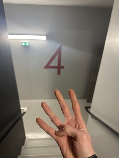 The image displays an indoor stairwell or landing area in Clichy, France. A large red numeral '4' is painted prominently on a light-grey wall in the mid-ground. Above and to the left of the '4', a horizontal fluorescent light fixture emits illumination, accompanied by two smaller green rectangular indicator lights below it. White stairs descend out of frame on both the left and right sides, flanked by a dark railing on the left and a white wall/railing with a dark handrail on the right. In the immediate foreground, a human left hand is positioned with its palm slightly upward and four fingers (index, middle, ring, and pinky) extended, while the thumb is bent inward, forming a visual representation of the number four. A dark denim cuff is visible at the wrist. The hand's gesture directly corresponds to the large '4' on the wall behind it.