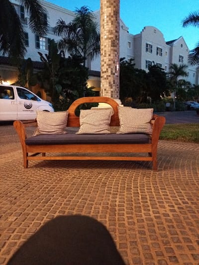 The image shows a wooden bench with three pillows on it. It is located on a patio made of small square tiles in front of a hotel building. The bench is facing the building.  There is a white van parked on the left side of the image.  The image was taken at coordinates -25.96, 32.605, which corresponds to a location in Maputo, Mozambique.  The image is taken from a low angle, and the viewer's shadow is cast over the tiled area in front of the bench.  The bench is empty and appears to be waiting for guests.
