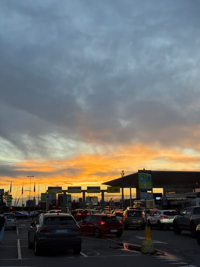 The image captures a view of an airport parking area at sunset in Orio al Serio, Italy. The sky is divided between dark, clouded skies on the left and orange and yellow sunset hues on the right. Rows of parking booths are visible, along with traffic signs and directional markers. Cars are lined up in the parking lanes, including a gray Audi Q3 and a red vehicle with the license plate "CR-017VC". A parking sign indicates "POSTI AUTO RISERVATI TERMINATION SPACES" and features a wheelchair icon.
