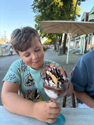 In Rostock, Germany, a young boy is seated at an outdoor cafe table, holding a large glass of ice cream. The ice cream is topped with whipped cream, chocolate sauce, and chocolate shavings. The boy wears a light blue t-shirt with a colorful, cartoonish design featuring monstrous figures. He is looking down at the ice cream with a slight smile. Beside him, a person wearing a blue shirt is also seated at the table. The background shows a treelined street with buildings, suggesting an urban setting, possibly a promenade or waterfront area given the presence of what appears to be a dock or harbor in the distance. An outdoor umbrella is visible behind the boy, providing shade. The sky is clear and blue.
