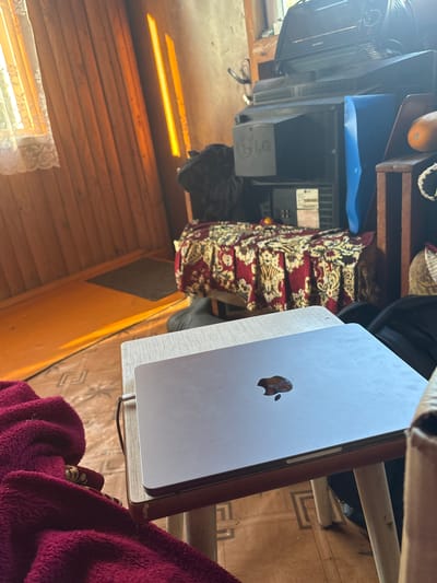 The image depicts an indoor scene, likely in a residential setting in Yurya, Russia, characterized by warm, natural light streaming in from a window, illuminating wooden walls and floor. A silver laptop with an Apple logo is placed on a small, light-colored table with visible wear. A dark red, fuzzy blanket is draped over the left side of the table and extends into the foreground. In the background, a collection of electronic equipment is stacked, including at least two CRT televisions, one bearing an LG logo, and other unidentifiable devices. A patterned red and gold cloth covers what appears to be a bed or sofa behind the electronics. The overall impression is one of a cluttered, lived-in space.