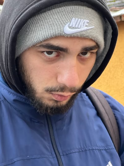A close-up, eye-level portrait captures a male individual with dark eyes, dark hair, a beard, and a mustache, looking directly forward. He is wearing a grey ribbed knit beanie with a white embroidered "NIKE" logo prominently displayed on the front. A dark-colored hooded sweatshirt is worn with the hood pulled over the beanie. Beneath this, a blue athletic-style jacket with a dark central zipper is visible. A dark backpack strap is seen over his right shoulder. The background is out of focus, revealing a light, indistinct surface to the left and a vivid yellow or ochre wall to the right. This scene is situated in Meistratzheim, France.