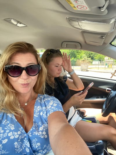 Two women are inside a car in Palje Brdo, Croatia. The woman in the foreground, with blonde hair and wearing a blue floral dress and sunglasses, is taking a selfie. The woman in the background, with blonde hair tied up and wearing a black top and white shorts, is looking at her phone and adjusting her hair. The car's interior is visible, including the dashboard, steering wheel, and roof lights. Outside the car, trees and a fence can be seen. The lighting suggests it is daytime.