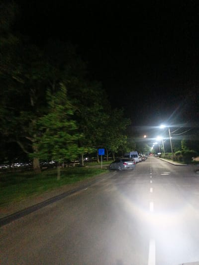 This image captures a night scene on a street in the city of Pivdenne, Ukraine. The perspective is from the driver's seat of a vehicle, with bright headlights illuminating the road ahead. The asphalt road is lined with white dashed markings and has a darker strip running down the center. On the left side of the road, a row of parked cars is visible, beyond which are trees and a grassy area. A blue parking sign is also present on the left. On the right side of the road, there are more trees and bushes, and several streetlights are casting bright illumination. In the distance, the road curves slightly and appears to extend into the darkness, with more streetlights visible. The overall atmosphere is dark, with the artificial lighting from streetlights and the vehicle's headlights creating strong contrasts.