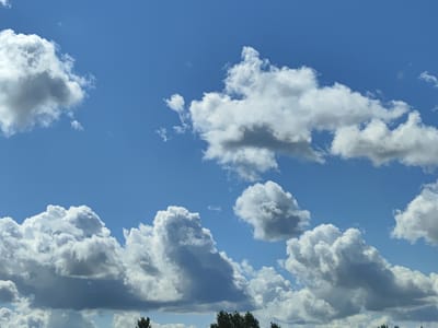 This is a wide-angle, eye-level photograph of a bright blue sky with several large, puffy, white cumulus clouds. The clouds vary in size and density, with some appearing almost entirely white and others showing darker grey undersides, suggesting depth and potential for precipitation. A thin strip of green foliage from trees is visible at the very bottom of the frame, indicating a ground-level perspective. The image captures a sunny day with clear visibility. The location is the municipality of Stoicini, Latvia.