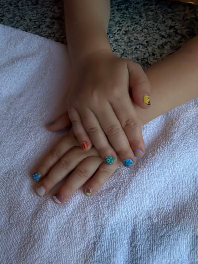 The image shows a close-up view of a child's hands with decorated fingernails resting on a white towel. The fingernails are adorned with various colorful designs, including what appear to be smiley faces, flowers, polka dots, and stripes. The background consists of a textured surface, possibly a granite countertop, and the white terrycloth towel. The lighting suggests an indoor setting. The image does not contain any information that can be used to determine its location in Urusovo, Russia.