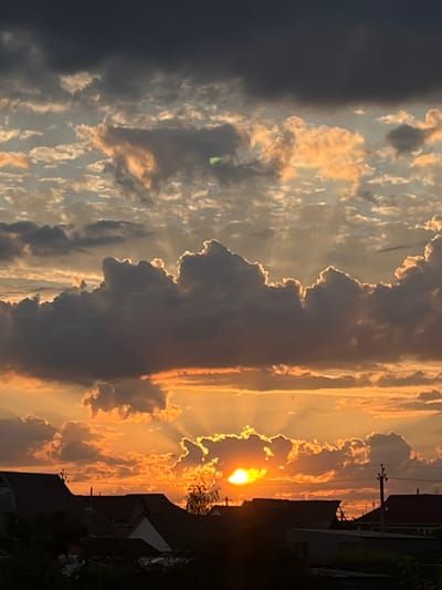 This is a photograph of a sunset over a residential area in Slobozia, Moldova. The sky is filled with dramatic clouds, some dark and heavy, others illuminated by the setting sun with hues of orange, yellow, and pink. Rays of light are visible emanating from behind the clouds. In the foreground, the silhouettes of rooftops and trees are visible, suggesting a suburban or rural setting within the city. A power pole with wires is also visible on the right side of the frame. There are no people or animals depicted in the image.