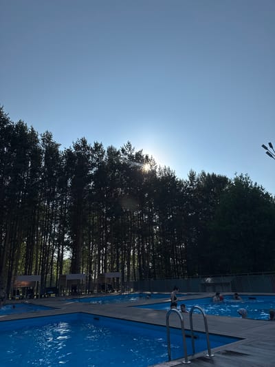 The image shows an outdoor swimming pool complex nestled amongst a dense forest of tall pine trees. The sun is partially obscured by the trees, creating lens flare and casting a bright glow across the sky. Several people are visible in the blue water of the pools, swimming and interacting. Wooden decking surrounds the pools, with some lounge chairs and covered cabanas visible in the background. The overall impression is a recreational area within a natural, wooded setting. The location is not specified as Berezovo, Russia.