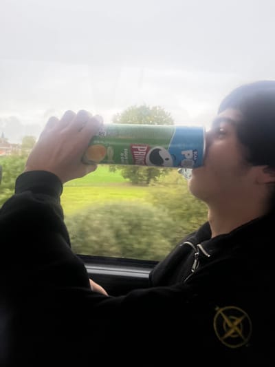 A male individual is depicted in Les Étangs, France, from the chest up, seated inside a vehicle. He is positioned facing left, holding a green and blue Pringles can labeled "Sour Cream & Onion" horizontally to his mouth, consuming its contents. The individual wears a dark, zippered hoodie, which features a circular yellow emblem with a cross-like design on the left chest area. Through the vehicle's window, an overcast sky is visible above a landscape of green fields and trees. In the distance to the left, a light-colored building with a red roof and a spire is partially observable.