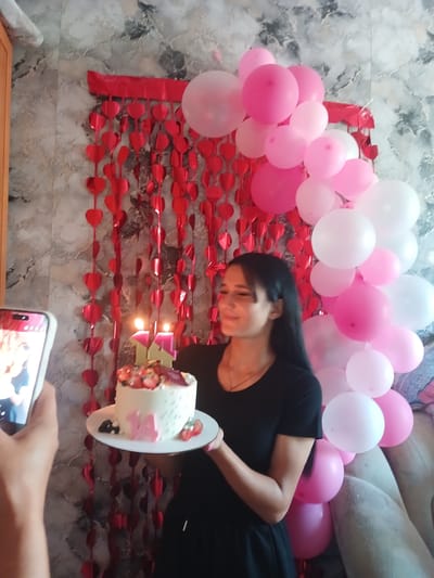 A young woman in a black dress stands in a room decorated with pink and white balloons and red heart garlands. She is holding a white cake topped with strawberries and has the number "14" candles on it. The woman is looking down at the cake with a smile. The background shows a textured wallpaper and a couch. The image appears to be taken indoors, likely in a residential setting. There is no information about the city of Chik, Russia.