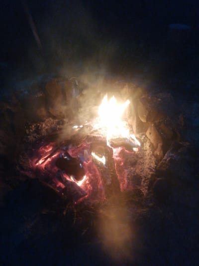 This is a close-up, low-light photograph of a campfire at night. The fire is burning brightly in the center of the image, with flames visible. Embers and glowing wood are also visible beneath the flames. The surrounding area is dark and appears to be covered in dirt or ash. There are some rocks or larger pieces of wood surrounding the fire pit. Smoke is rising from the fire and is illuminated by the flames. The overall impression is one of a rustic outdoor setting. The image does not contain any people, or any discernible elements that would place it specifically in Шарпілаўскі сельскі Савет, Belarus.