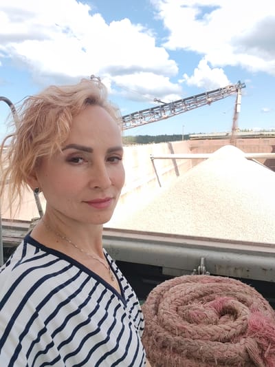 A blonde woman with short, styled hair and light makeup is looking towards the camera, with her head turned slightly to the left. She is wearing a black and white striped V-neck top and a delicate silver necklace. Her right ear is adorned with a circular earring featuring a dark stone. In the background, a large conveyor belt system is visible, transporting a light-colored granular material. The material is piled high in a large, outdoor industrial setting, possibly a port or quarry. The sky above is blue with scattered white clouds. In the foreground, a thick, coiled rope, predominantly pinkish-brown in color, is prominent. The scene appears to be in an industrial area of Kvartsitny, Russia.