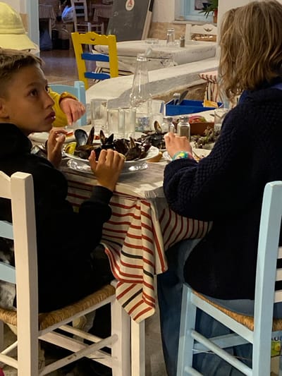 The image portrays an indoor dining scene at a restaurant in Megalochori, Greece. A young male with short brown hair, wearing a black hooded top, is seated on the left side of a table, looking towards the left. His hands are positioned near a white platter laden with cooked mussels and lemon wedges, which occupies the center of the table. A second individual, with long, wavy blonde hair, dressed in a dark blue knitted top and blue jeans, is seated opposite the first, with their back to the camera; a colorful bracelet is visible on their right wrist. A third person's hand, wearing a yellow sleeve, is partially visible behind the young male.

The table is covered with a white tablecloth, overlaid with a decorative red, white, and black striped fabric. Other items on the table include multiple clear glass pitchers, smaller drinking glasses, salt and pepper shakers, a blue holder containing utensils, and a small dish containing what appears to be grapes. The chairs are wooden with woven straw seats, in white and light blue colors. In the blurred background, the restaurant's interior features white walls, arched architectural elements, additional tables and chairs, a yellow and blue chair, and a dark board displaying a white 'A' logo.