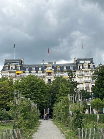 The image displays the Beau-Rivage Palace hotel in Lausanne, Switzerland, viewed from a park on an overcast day. The grand, multi-story building occupies the upper portion of the frame, featuring a light-colored facade, a dark mansard roof with numerous dormer windows, and multiple balconies, several adorned with extended yellow awnings. Two flagpoles on the roof prominently display a green flag on the left and a red flag on the right.

The foreground is a park setting with a light-colored gravel path leading towards the hotel. The path is bordered on both sides by rows of green plants, some supported by vertical wooden trellises. A low wooden fence is visible along the bottom left edge. Dense green deciduous trees partially obscure the lower levels of the building and define the mid-ground. Approximately three to four individuals are visible standing in a small cluster on the gravel path near the base of the trees in the mid-ground. The sky above is entirely covered with dark grey, overcast clouds. Faint outlines of parked or moving vehicles are discernible behind trees on the far right.