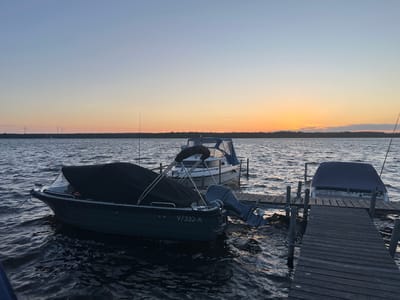 The image depicts a tranquil scene at sunset on the Schwielowsee in Schwielowsee, Germany. The sky exhibits a gradient of pale blue to warm orange as the sun dips below the horizon, casting a soft glow over the water. A wooden pier extends into the lake, supporting several boats. In the foreground, a dark-colored boat is moored, its bow pointed towards the viewer and covered with a black tarp. An outboard motor is visible at its stern. Adjacent to this boat, another vessel, larger and white, is also docked and covered. Further down the pier, a third boat is partially visible, also covered. In the distance, across the lake, a tree-lined shore is silhouetted against the sunset, with several wind turbines dotting the landscape. The water is gently rippled, reflecting the colors of the sky. There are no people visible in the image. The overall atmosphere is peaceful and serene.