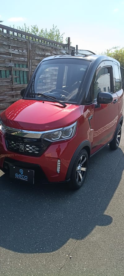 This media file shows a red electric car, specifically a Geco E-Automobile, parked on an asphalt surface, likely a street or parking lot in Mühlhausen, Germany. The car is viewed from the front-right angle, with the driver's side door open and the interior visible. The sun is shining, casting a shadow of a person onto the asphalt, indicating it's daytime. The background features a wooden fence and some greenery, suggesting an outdoor setting. The car has a sleek design with distinctive headlights and a prominent grille. The "Geco E-AUTOMOBILE" logo is visible on the rear bumper. The image is taken from a slightly low angle, emphasizing the car's presence. The overall scene appears to be a casual one, perhaps a demonstration or personal vehicle.