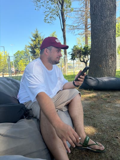 A man wearing a maroon baseball cap, a white t-shirt, and tan shorts is sitting on a large gray beanbag chair outdoors. He is holding a black smartphone in his right hand and appears to be looking at it. His left hand is resting on his leg. He is wearing dark green flip-flops. Behind him, to his left, are trees and a metal fence. To his right is a large tree trunk and a black beanbag chair. The ground is covered in grass and dry leaves. The sky is clear and blue. The scene is likely in a park or outdoor recreational area, possibly within the city of Babyshira, Abkhazia, given the mention of this location.