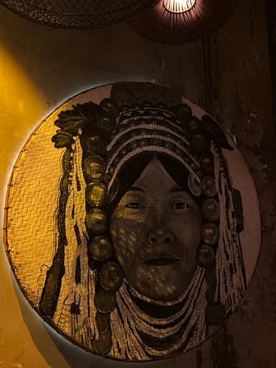 An indoor shot shows an Indonesian artwork hanging on a wall. The artwork is round and made of woven material. A face is painted onto the round artwork, featuring eyes, a nose, and lips. Above the face, there is another design. The artwork is positioned with the face in the center, facing forward. To the left of the main artwork, there is another, similar object made of woven material. The setting appears to be in Pererenan, Indonesia.