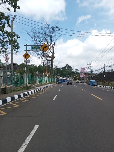 This image depicts a daytime street scene in Boommlog, Bhutan, under a partly cloudy sky. An asphalt road, marked with white solid and dashed lines separating lanes and a yellow dashed line further right, extends toward the horizon. The road features black and white striped curbs along both sides, with yellow no-parking markings visible on the pavement adjacent to the left curb.
On the left side of the road, a green metal fence runs parallel to the street, behind which are visible parts of buildings and green foliage. A utility pole with multiple power lines extending across the frame is present on the far left. A metal signpost stands prominently on the left, displaying a green rectangular sign with white text "Jl. Raya Puncak" and two yellow diamond-shaped signs, each featuring a black arrow indicating a sharp curve to the right. Below these, a white rectangular sign reads "Tikungan Tajam Ke Kanan." A circular red sign with a diagonal line through a 'P', indicating no parking, is also attached to the signpost.
Vehicular traffic includes several blue microbuses or vans, a dark SUV, and a light-colored truck, all traveling away from the viewer in the left lanes. Another blue microbus is parked on the right shoulder of the road. Overhead, numerous power lines traverse the scene. Trees with varying foliage, including one large deciduous tree with mostly bare branches near the signs, line the roadside. In the distance on the right, a large billboard with pink and white text and a decorative archway structure are discernible.
