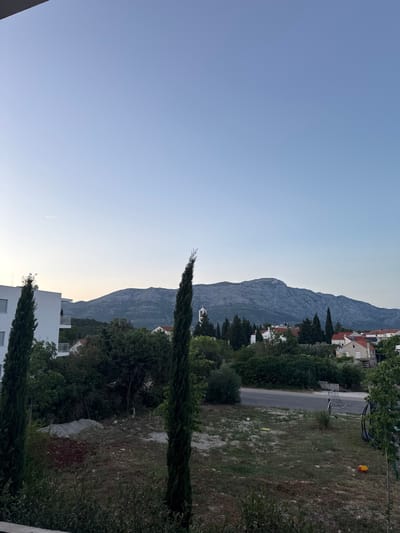 This image captures an outdoor scene in Grad Korčula, Croatia, during what appears to be either early morning or late evening, characterized by soft, diffused light and a clear, pale blue sky.
The upper portion of the frame is dominated by a clear sky, transitioning from a light blue at the top to a nearly white-blue near the horizon line. A faint white contrail from an aircraft is visible in the upper left quadrant.
In the background, a large, rugged mountain range with a distinct grey, rocky texture stretches horizontally across the frame. Below the mountain, the landscape is dotted with numerous green trees and clusters of traditional buildings, primarily houses with orange-red tiled roofs. A slender white church steeple with a pointed roof is discernible on the left side of the mountain base.
The midground features a mix of dense green foliage and architectural elements. On the far left, a modern, multi-story white building with balconies is partially visible. Two tall, slender cypress trees stand prominently, one on the left foreground and another on the right, framing the view towards the mountains and the distant settlement. Various other trees and bushes with dark green leaves fill the space between the foreground and the mountain range.
The foreground consists of an uneven plot of undeveloped land covered with sparse, dry grass, patches of reddish-brown dirt, and scattered light-colored rocks. A paved road is visible running horizontally through the mid-to-foreground, with more vegetation on the far side. Specific objects on the foreground plot include a small white soccer ball, a lightweight white and black mini soccer goal, and a yellow plastic toy. No people or animals are visible in the scene.