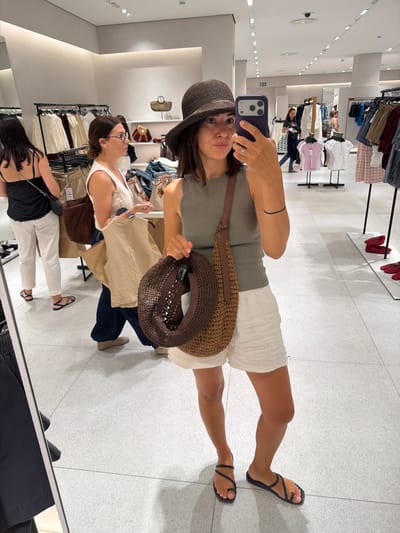 A woman is taking a mirror selfie in a clothing store. She is wearing a grey sleeveless top, white shorts, a dark bucket hat, and black sandals. She is holding a woven straw bag over her shoulder and another straw hat in her hand. In the background, other shoppers are browsing clothes on racks. The store appears to be a modern retail establishment with bright lighting and a tiled floor. The location is identified as Alboraia / Alboraya, Spain.