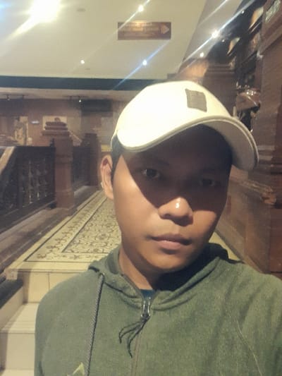 This image depicts a male individual captured in a selfie format, situated within an indoor architectural space in Glantengan, Indonesia. The subject wears a dark green zipped hoodie and a light-colored baseball cap. Direct overhead lighting causes a glare on the forehead and partially shadows the upper facial features. The background features a hallway or corridor characterized by dark, carved wooden or stone structures, including railings on the left, indicative of traditional Indonesian design. The floor is covered by a patterned runner with light, decorative motifs. Steps are visible on the lower left. A rectangular brown sign with an arrow pointing right is mounted on the upper left wall. The scene is well-illuminated by recessed ceiling lights and appears devoid of other individuals.