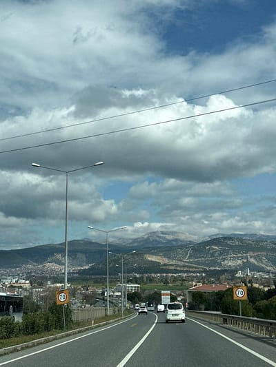The image displays a daytime view from a vehicle on a multi-lane highway leading towards the city of Menteşe, Turkey. The sky is partially covered by large, grey and white cumulus clouds, with several patches of blue sky visible. Two thin, dark power lines cross the upper portion of the frame.
In the foreground, a paved road with white solid lane markings and a yellow dashed line extends into the distance, curving slightly to the right. A tall, grey streetlight pole with a dual horizontal lamp fixture is prominent on the left side of the road. A yellow circular speed limit sign, featuring a black border and the number '70' in black, is positioned on a pole to the right of the road. Further down the road, a series of similar streetlights recede from view.
Several vehicles are observed on the road, all moving away from the camera. A white panel van occupies the right lane, followed by a white SUV. A dark car and another white vehicle are visible further down. On the far left, a portion of a large, dark vehicle, possibly a bus, is partially obscured behind a concrete barrier that lines the left side of the highway. A metal guardrail runs along the right side.
The midground reveals the urban landscape of Menteşe, characterized by various buildings and interspersed green vegetation. On a hillside to the right, a large quarry or excavation site with exposed rock faces is visible. The background is dominated by a mountain range, with the most distant peaks appearing distinctly snow-capped, while closer mountains are covered with trees and sparse vegetation.