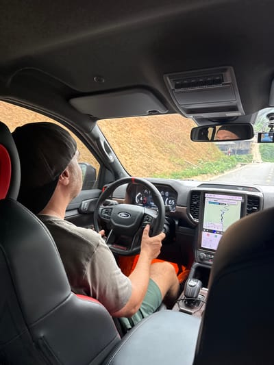 The media file captures the interior of a vehicle from a high angle, looking forward from the rear passenger side. A male driver, visible from behind, wears a black cap, a grey t-shirt, and green shorts. His left hand rests on the steering wheel, and his reflection is faintly visible in the rearview mirror.
The vehicle's interior features a large, vertical infotainment screen displaying a navigation map with a route line and an elevation profile, showing the time "13:01" and a temperature of "22.5°". Below the screen, the dashboard and center console are visible, including a rotary gear shifter with "TRX" stitched in red on its boot. A small screen behind the steering wheel shows the speed as "65". An overhead console above the rearview mirror contains six auxiliary switches labeled "AUX 1" through "AUX 6". A small camera or device is mounted on the windshield. The steering wheel, however, clearly features a "Ford" logo and "Raptor" text, creating conflicting brand identifiers within the vehicle's cabin which otherwise strongly suggests a Ram TRX due to the large screen and shifter design.
Outside the car, a road is visible ahead, bordered by green and brown vegetation, consistent with a rural or semi-rural landscape. The scene indicates bright daylight and clear weather. The overall activity is driving and navigation, likely within Minh Sơn Commune, Vietnam, given the contextual information. No specific emotions are discernible from the partially obscured driver.