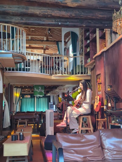 The image depicts a dimly lit indoor venue, possibly a cafe or bar, in Dali City, China, characterized by its rustic, multi-level wooden architecture. A young woman is seated on a wooden stool on a raised stage, playing an acoustic guitar. She is dressed in a white long-sleeved top and matching white pants. A microphone is positioned in front of her. Behind her, leaning against a wooden wall, is another acoustic guitar. To the right of the stage, musical equipment, including a keyboard and speakers, are visible.

The venue features multiple levels with wooden railings and stairs, creating an open, loft-like atmosphere. In the background, teal curtains and some tables and chairs are discernible, suggesting a seating area. The decor includes bookshelves filled with books and some decorative items like a chandelier. The overall ambiance appears to be intimate and designed for live performances.