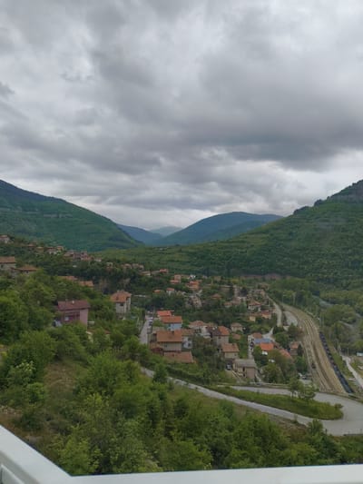 A high-angle view captures the Bulgarian village of Bov, nestled in a valley surrounded by green, tree-covered mountains under an overcast sky. The village is characterized by closely spaced houses with red-tiled roofs. A winding road and railway tracks run through the village. A train is visible on the tracks, suggesting possible rail transport. The overall setting suggests a rural or semi-rural environment.