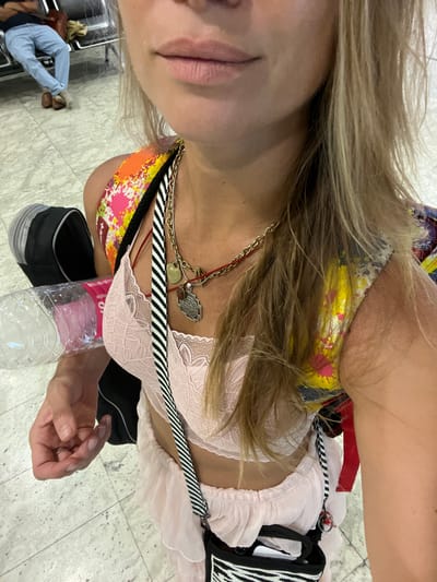 The image shows a close-up of a person's upper body, with a focus on their clothing and accessories. The subject has blonde hair that partially covers their face. They are wearing a pink lace bra, and a colorful patterned bikini top is visible underneath. A black and white striped strap crosses over the bra. Around their neck, they have multiple necklaces, including a gold chain with a pendant and a red string with a charm. In the foreground, a partially filled plastic water bottle rests against the subject's chest.
In the background, the scene appears to be an indoor public space with a tiled floor. To the left of the frame, two people are partially visible in the distance. One is wearing light blue pants and appears to be sitting or crouching. The other person is mostly obscured. The overall impression is that the photo was taken in a casual setting, possibly an airport or a public waiting area, given the flooring and the presence of other people. There is no indication of the time of day or weather conditions.