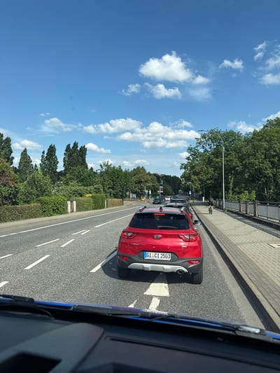 The image shows a roadside scene in Giessen, Germany on a sunny day. The perspective is from inside a vehicle, looking out towards a red Kia SUV driving in the same direction. The SUV has a German license plate that reads "GI CI 2503". The road has multiple lanes, and there is traffic visible in the distance, including other cars and a truck.

To the left of the road, there is a sidewalk and a grassy area with trees and bushes. A street light with a traffic signal is visible, showing a green light, indicating traffic is moving. The sky is blue with scattered white clouds, suggesting fair weather. The sun appears to be high in the sky, indicating it is likely daytime, possibly late morning or afternoon.

The scene appears to be a typical urban or suburban environment. There are no people prominently featured in the image, though a person is partially visible on a bicycle on the sidewalk. The overall mood is calm and uneventful, characteristic of everyday traffic. The image provides a snapshot of a journey through a German city on a pleasant day.