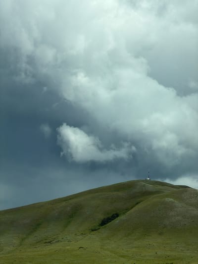 A solitary communications tower stands atop a grassy, undulating hill, its white structure contrasting with the overcast, dramatic sky. The hill is covered in a dense, short green grass, with subtle shadows indicating the rolling contours of the landscape. The sky is filled with heavy, gray clouds that create a somber and somewhat ominous atmosphere. The scene appears to be a rural or remote location, possibly on the outskirts of Верхнеозёрное, Russia, given the context provided, though no explicit urban elements are visible in the image. There are no people or animals present, and the focus is solely on the natural landscape and the man-made structure.